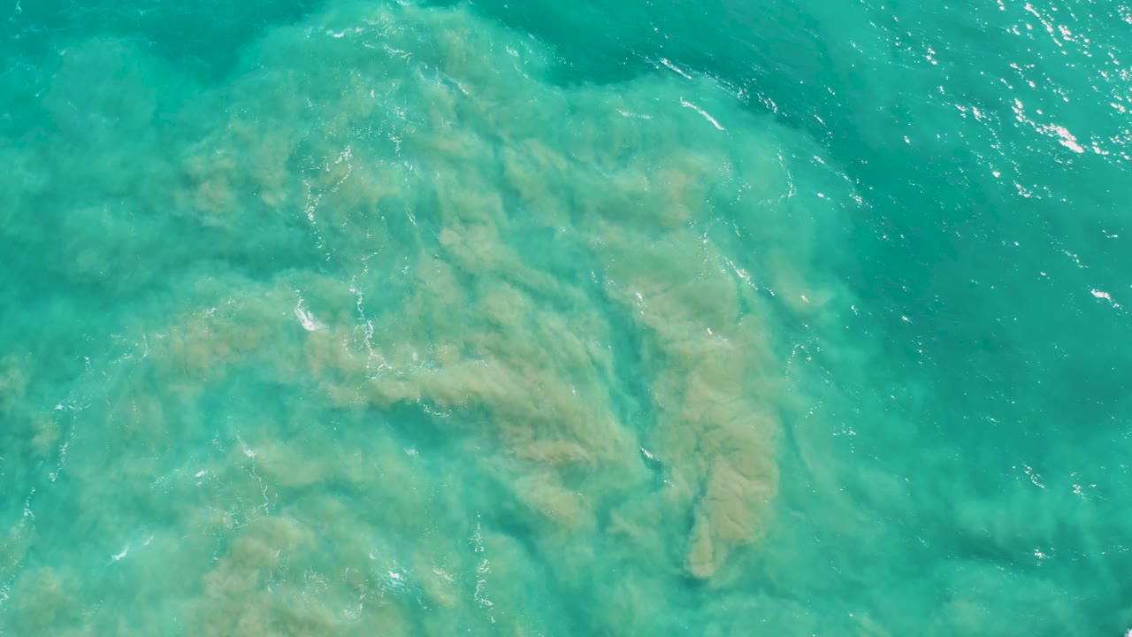 Clear blue-green water in San Jose Del Cabo covers a pale sandy ocean floor with rock shapes beneath and small surface ripples, no visible people.