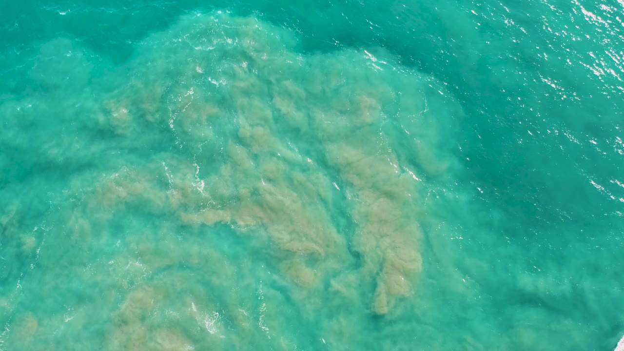 Aerial view of San Jose Del Cabo turquoise ocean with a broad submerged rock patch under the surface and brighter sun flecks near the upper right, no visible people.