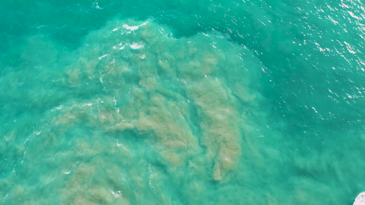 Overhead San Jose Del Cabo water view with sandy tan formations under turquoise ripples and a small white foam mark near the lower edge, no visible people.
