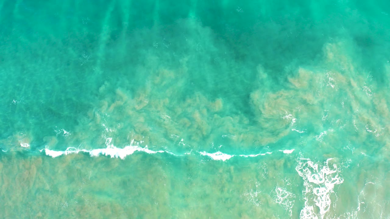 Top-down view of turquoise ocean water in San Jose del Cabo with pale seabed visible and a thin white wave crest crossing the lower frame, no visible people.
