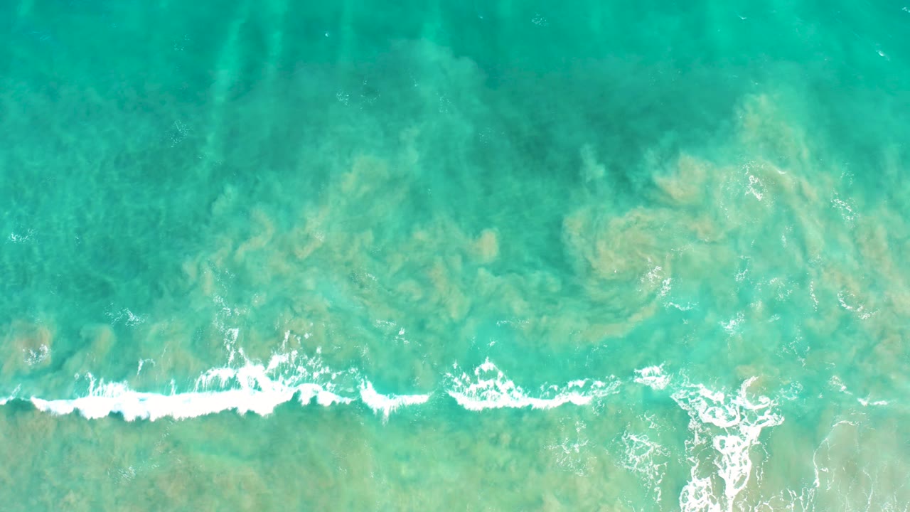 Top-down view of clear green-blue ocean in San Jose del Cabo as a broken white surf line stretches across the frame with tan seabed texture below, no visible people.