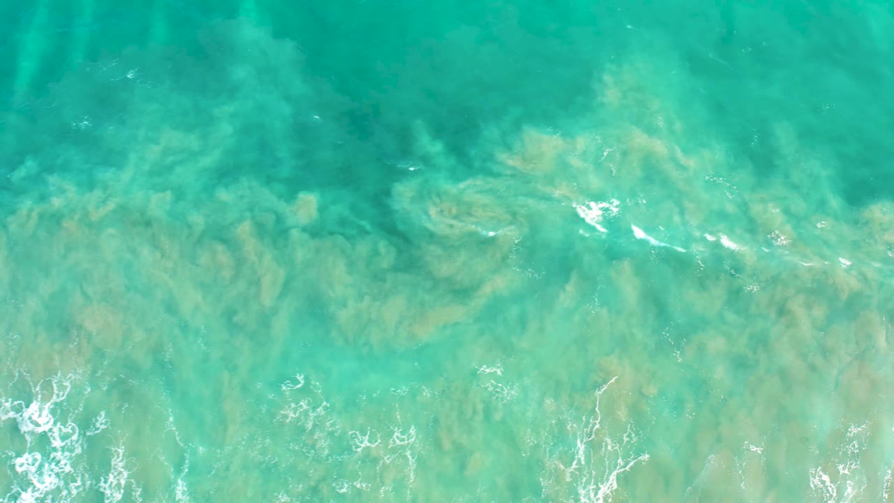 Aerial view of clear turquoise ocean in San Jose Del Cabo, with sandy seabed patterns visible beneath the surface and white foam gathering along the lower frame, no visible people.