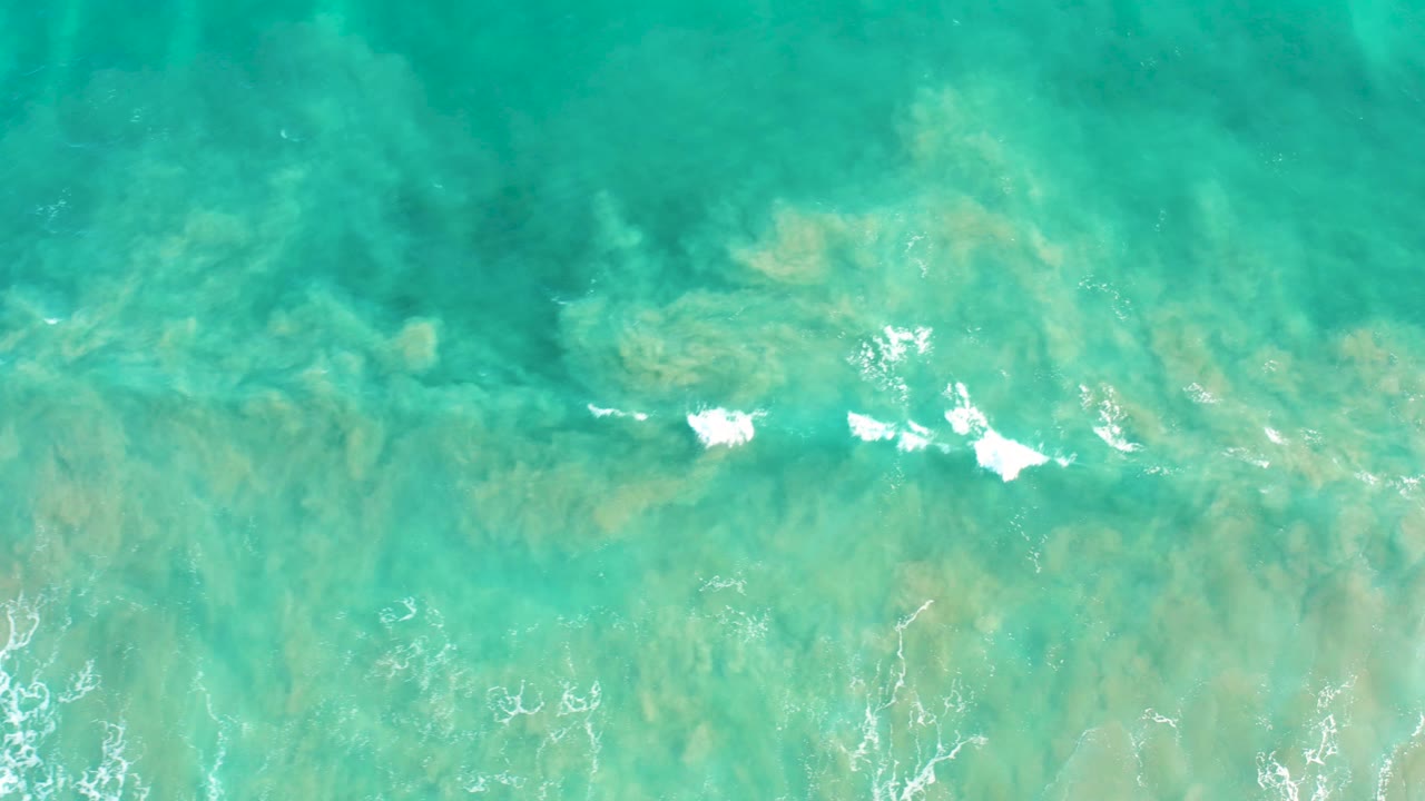 Top-down San Jose Del Cabo ocean frame with pale seabed swirls under turquoise water and broken white foam streaks crossing the lower portion, no visible people.