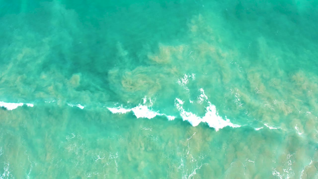 Aerial frame in San Jose Del Cabo with a more defined white wave line cutting across shallow turquoise water and the seabed still visible below, no visible people.