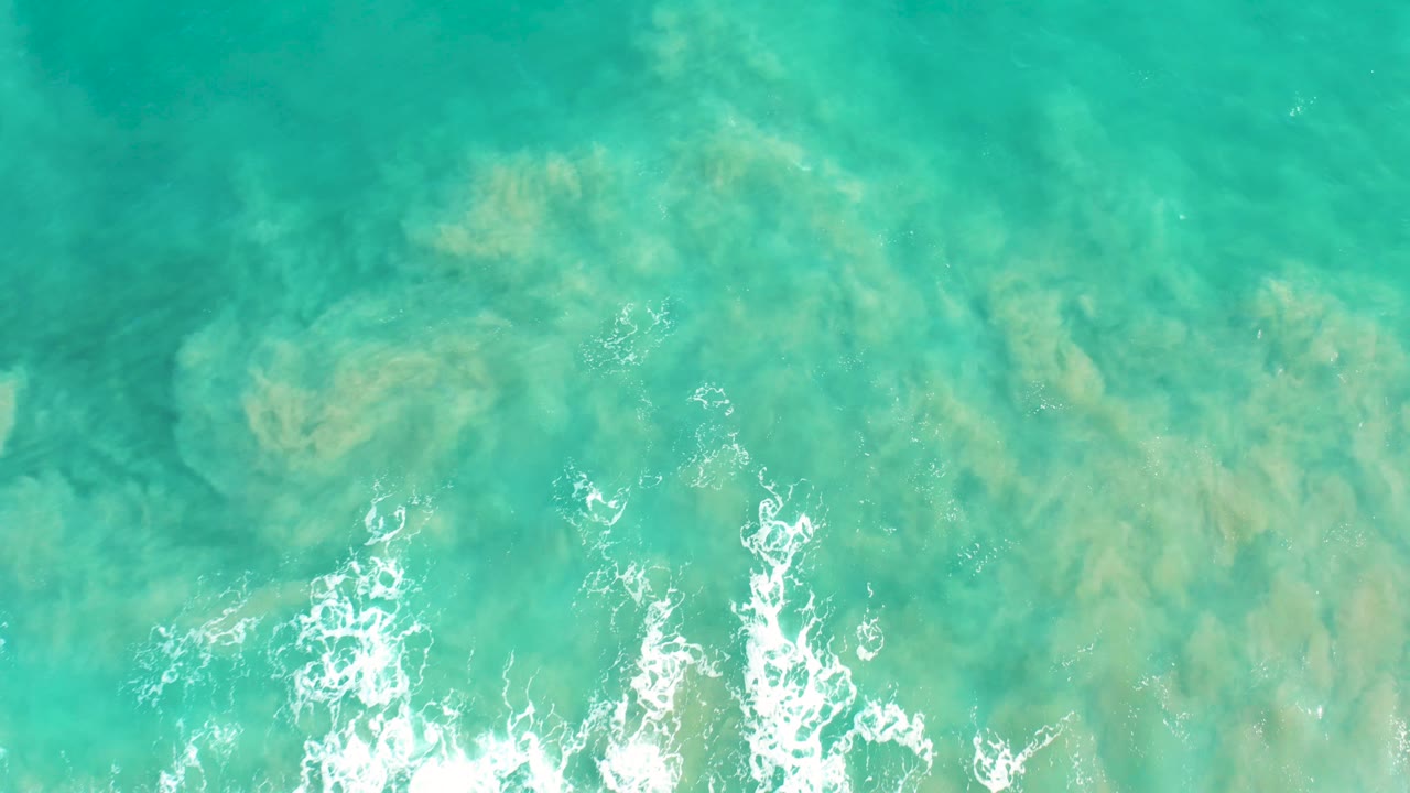 Top-down view of shallow turquoise ocean in San Jose Del Cabo, with pale seabed shapes under the water and white foam at the bottom edge, no visible people.