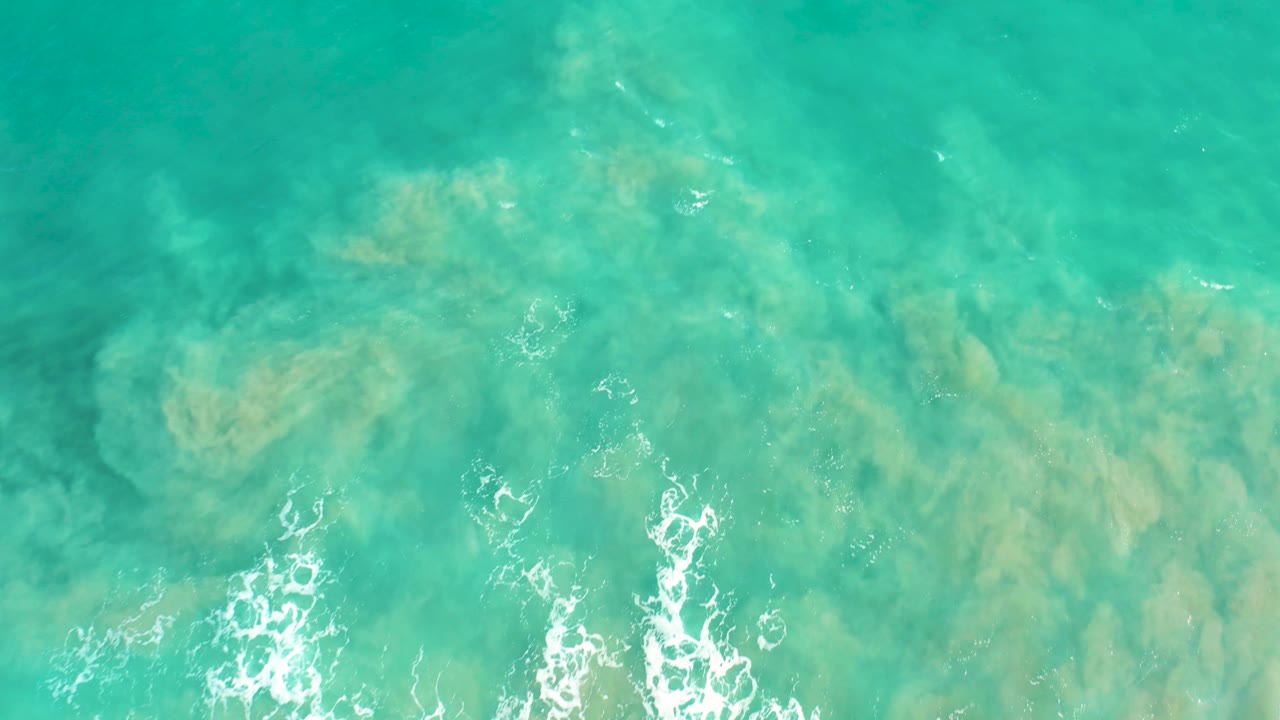 Aerial view of San Jose Del Cabo turquoise water with fading foam near the lower left and broad sandy seabed patterns visible beneath, no visible people.