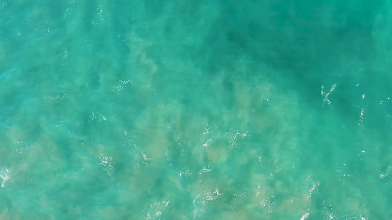 Clear blue-green ocean surface in San Jose Del Cabo with small white foam marks drifting over a visible sandy bottom and no visible people.