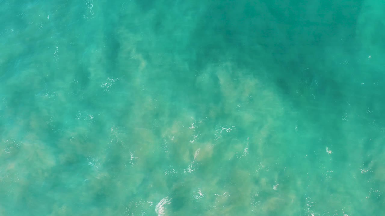 Transparent turquoise shallows in San Jose Del Cabo reveal pale sand patterns beneath the water, with scattered foam specks and no visible people.