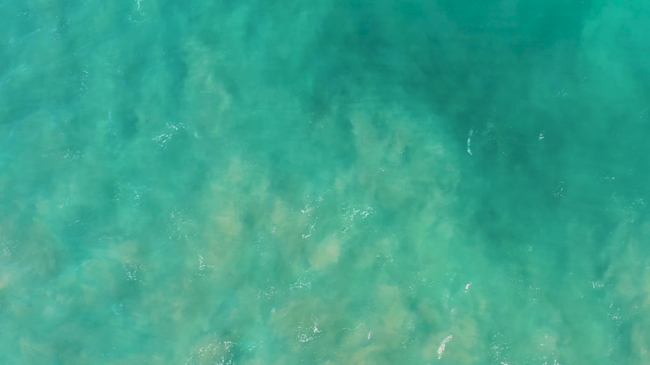 Top-down ocean view in San Jose Del Cabo shows blue-green water over a mottled sandy bottom, with faint white bubbles on the surface and no visible people.