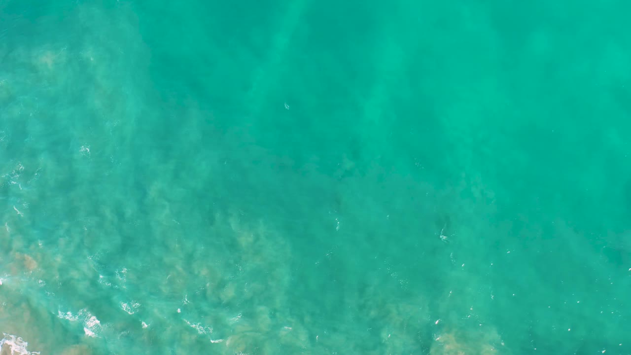 Clear teal water covers a mottled pale seabed in San Jose del Cabo, with faint white foam along the lower edge and no visible people.