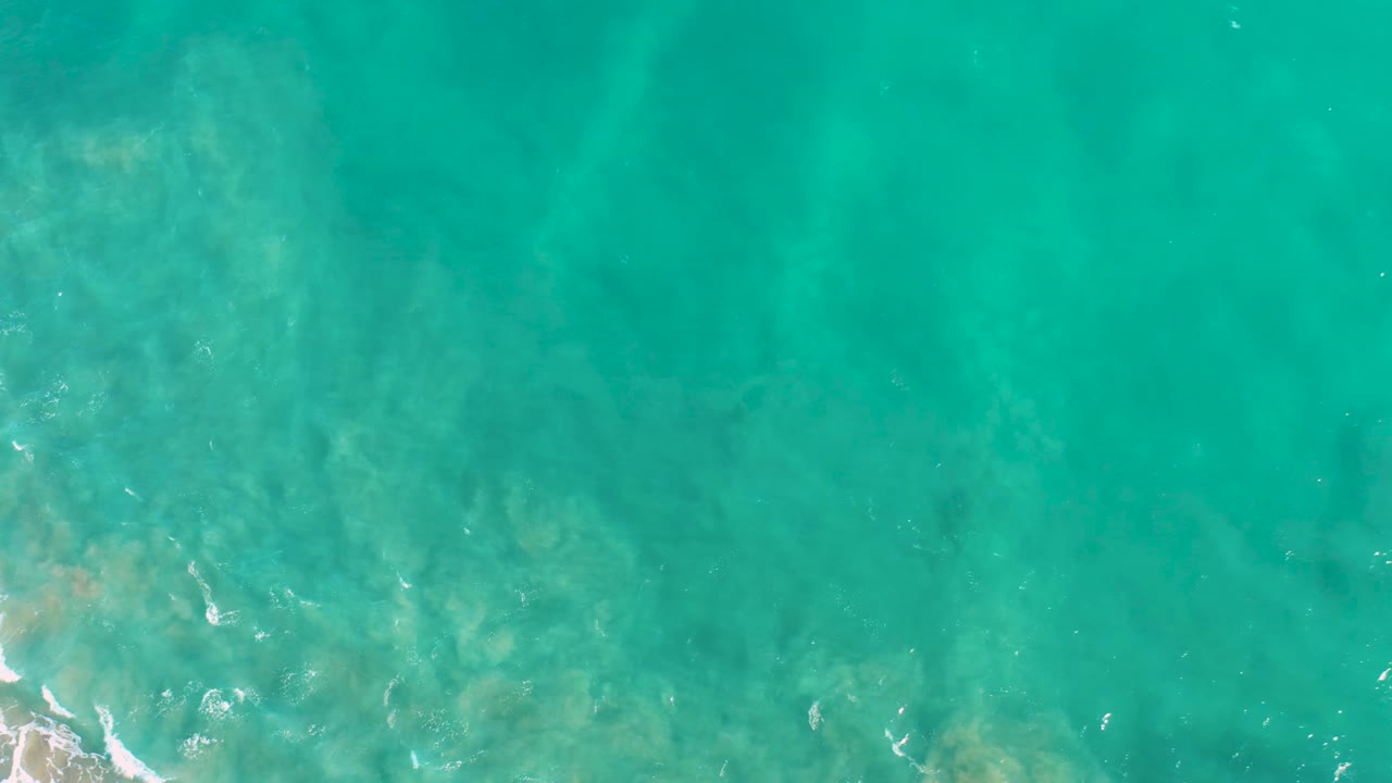 A top-down San Jose del Cabo ocean frame shows turquoise water, hazy sand color below, and scattered foam traces near the left side with no visible people.