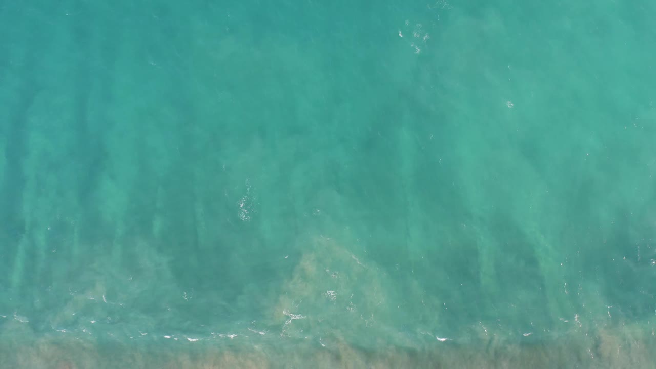 Top-down San Jose del Cabo ocean frame showing clear turquoise water, darker underwater bands, a thin broken foam line near the bottom, and no visible people.