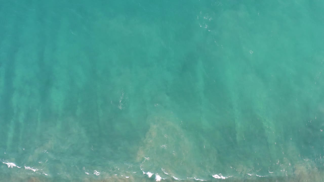 Aerial view over San Jose del Cabo turquoise sea with pale submerged rock or coral shapes, scattered white foam flecks, soft surface ripples, and no visible people.