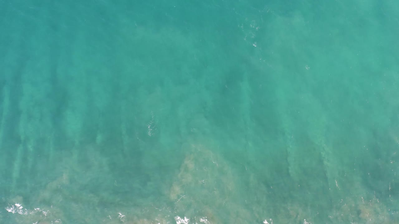 Top-down San Jose del Cabo water frame with turquoise surface color, darker vertical underwater streaks, pale seabed patches near the bottom edge, foam at the edge, and no visible people.