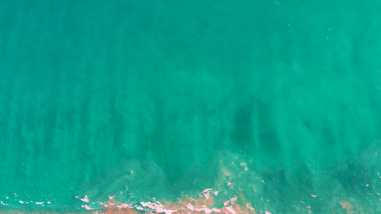 Aerial frame over San Jose Del Cabo water with darker sand streaks under the turquoise surface, a thin foam trace at the bottom, and no visible people.
