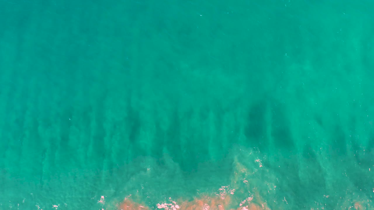 Aerial view in San Jose Del Cabo showing turquoise water, vertical-looking underwater bands, muted sand beneath the surface, and no visible people.