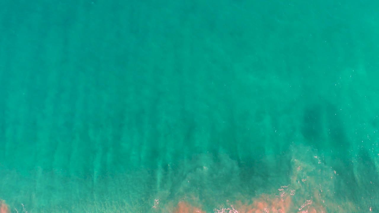 Top-down view of shallow turquoise water in San Jose del Cabo with vertical dark seabed patterns, pale tan areas near the bottom edge, and no visible people.