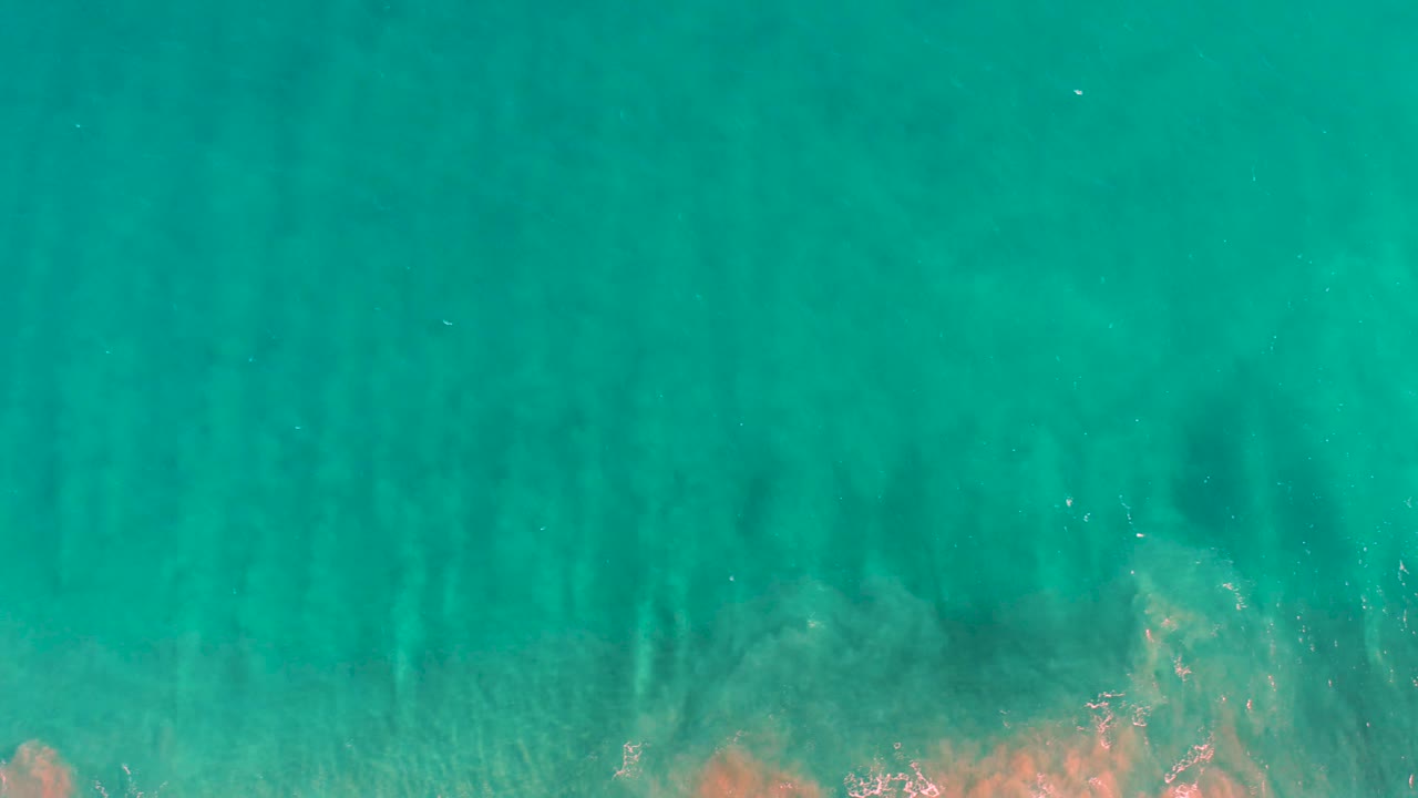 Aerial view over San Jose del Cabo shallow sea with turquoise water, soft vertical seabed streaks, and light tan patches touching the lower frame, with no visible people.