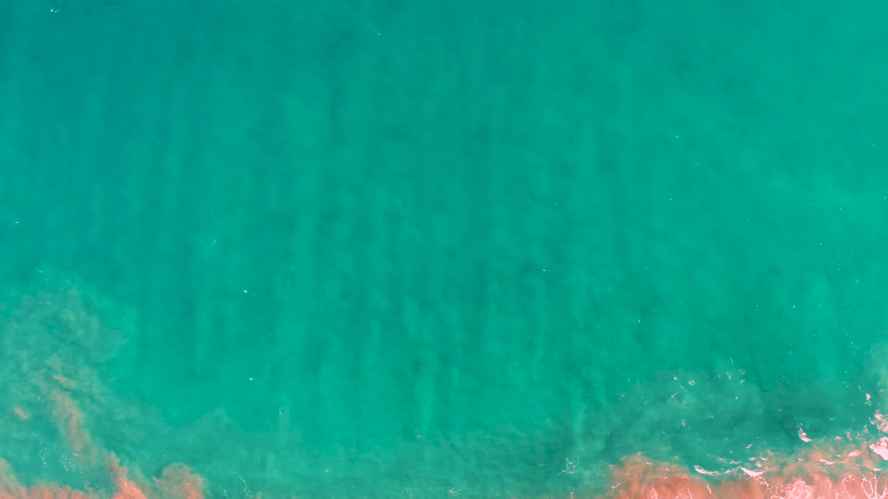 Turquoise shallow sea in San Jose del Cabo shows darker underwater ridge lines with a small orange-brown rocky or coral patch near the lower edge and no visible people.