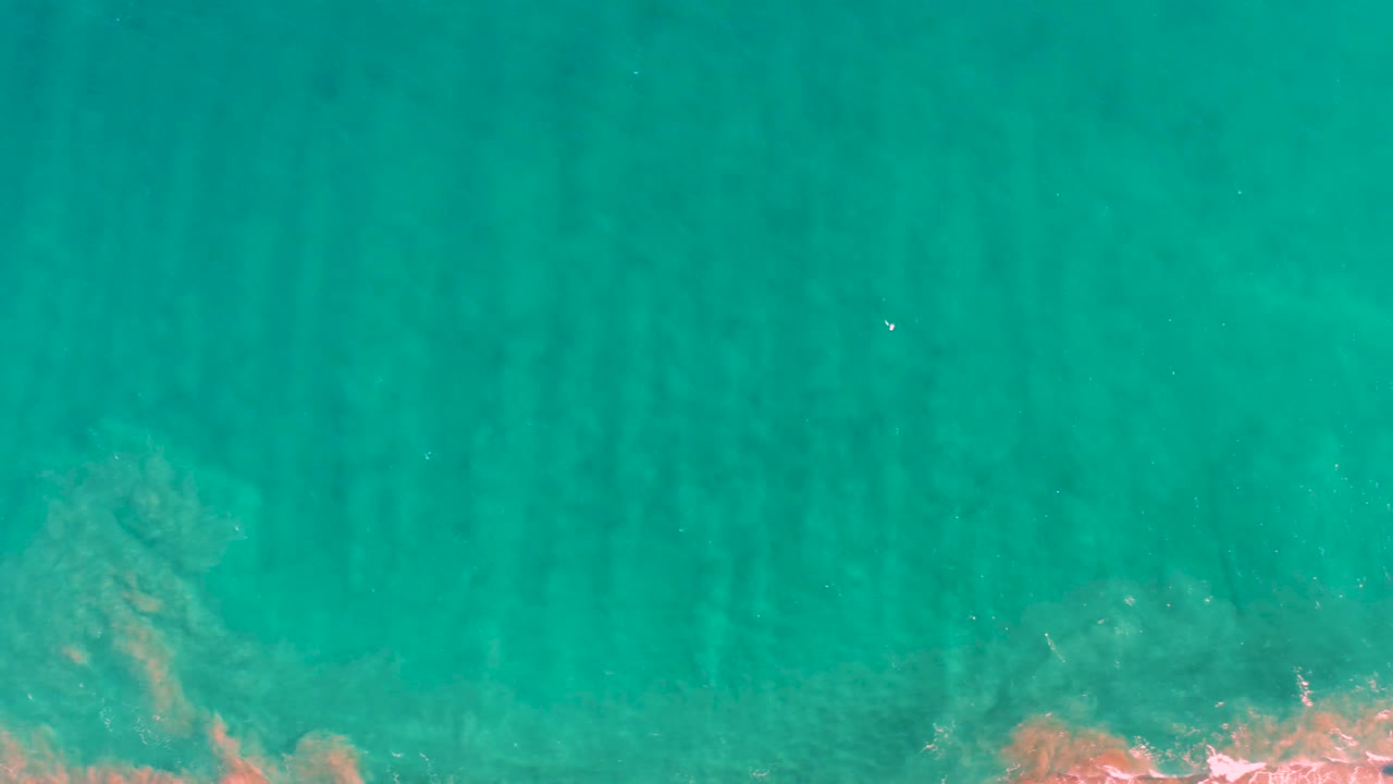 Clear turquoise water over shallow sand ridges in San Jose del Cabo, with a pale orange rocky or coral area touching the lower frame edge and no visible people.