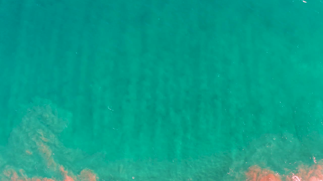Overhead San Jose del Cabo water view with striped underwater sand ridges, teal color, and orange-brown rocky or coral texture along the bottom border, with no visible people.