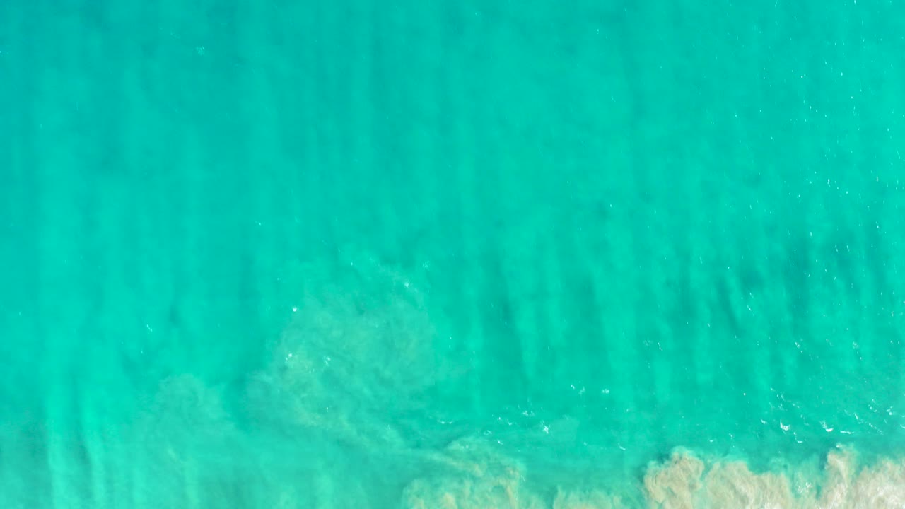 Top-down view of turquoise shallow water in San Jose del Cabo with faint dark seabed streaks and a thin line of white foam entering near the lower edge, no visible people.