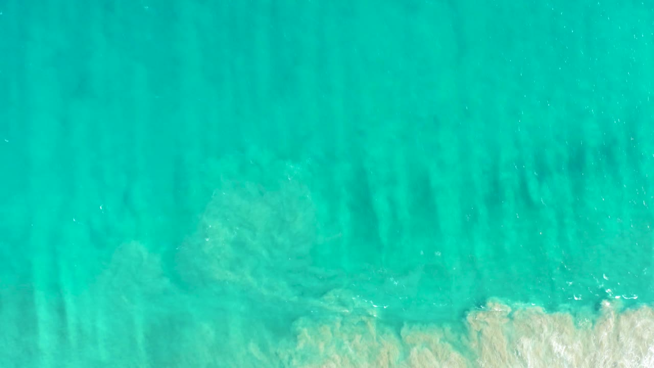 Aerial view of San Jose del Cabo turquoise shallows with darker seabed streaks and a small brighter patch of breaking white surf near the lower right, no visible people.