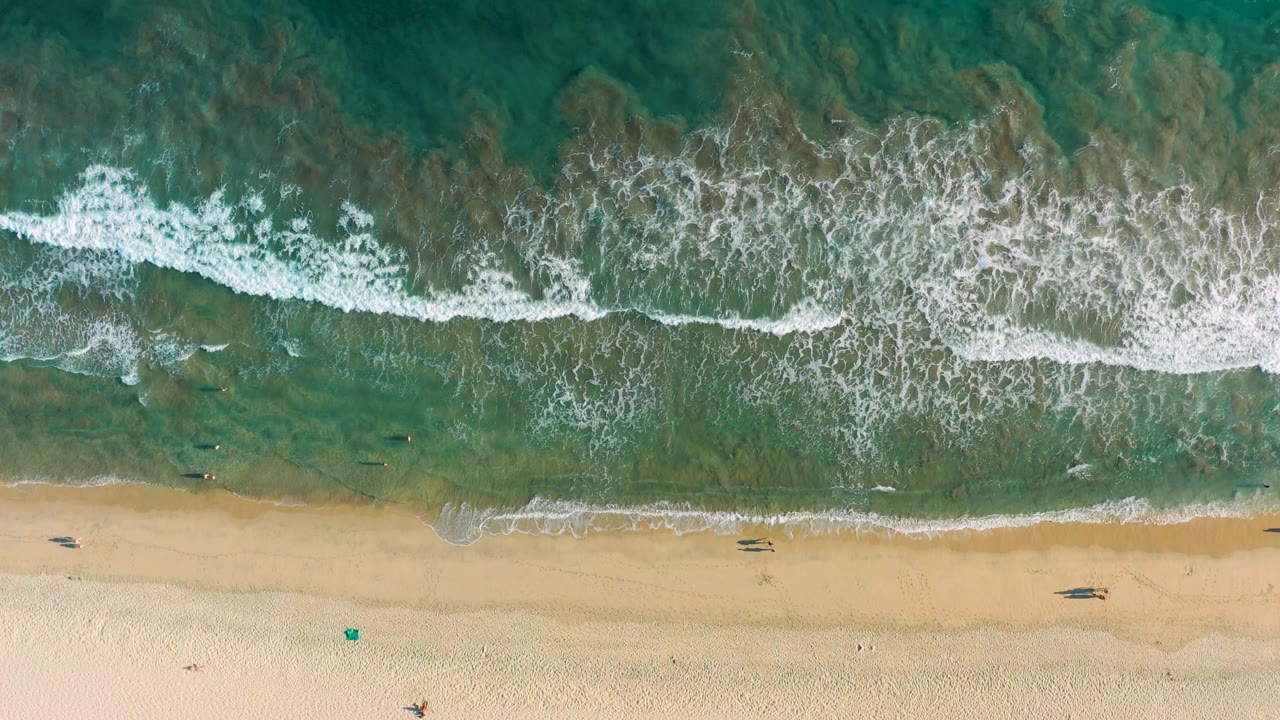 Overhead San Jose Del Cabo shoreline with turquoise and green water, broken foam near the upper half, and a mostly open sandy beach with tiny figures near the waterline.
