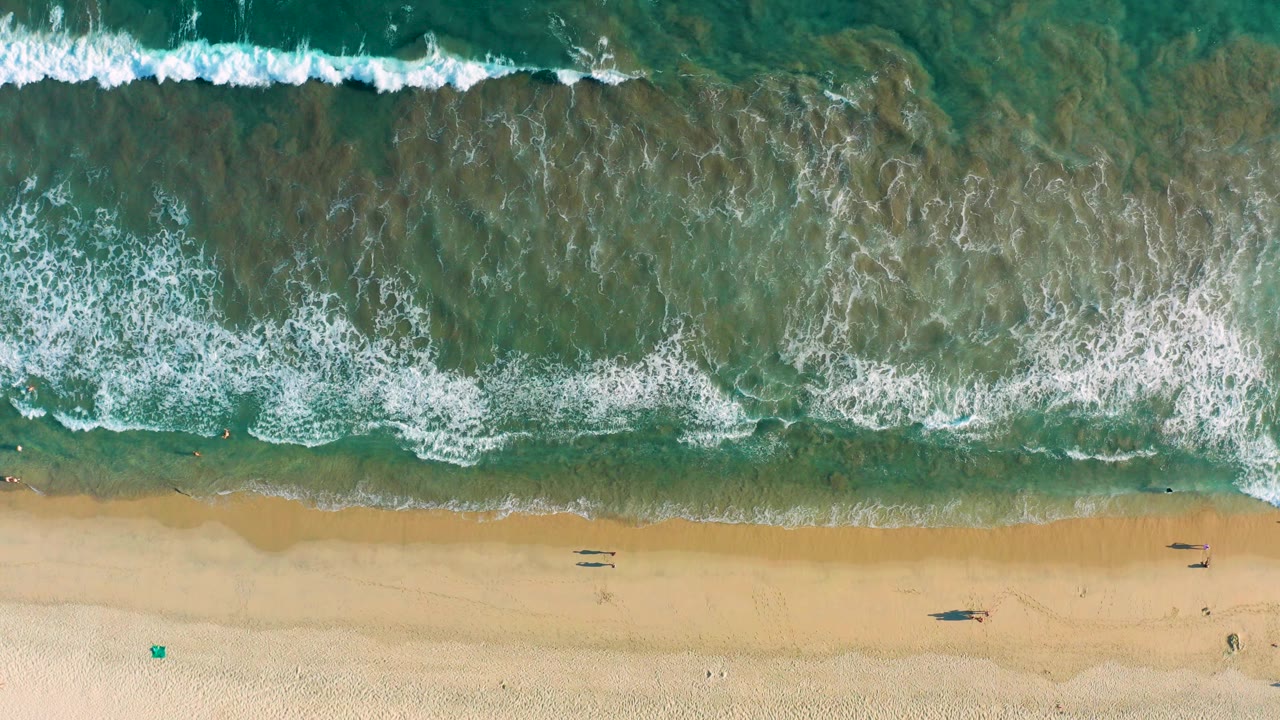 Top-down view of San Jose del Cabo surf, with turquoise water and white foam approaching a wide strip of pale sand.