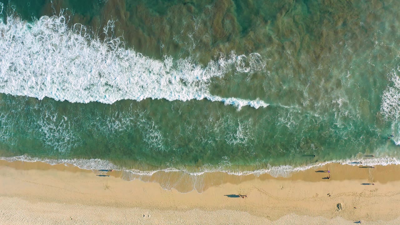 Straight-down view of San Jose Del Cabo beach with churning white foam across green-blue water and a thin tan shoreline below, with no visible people.
