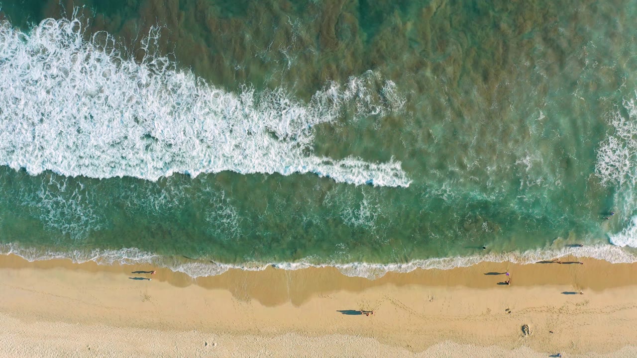 Wave foam reaches closer to the sandy beach from above.