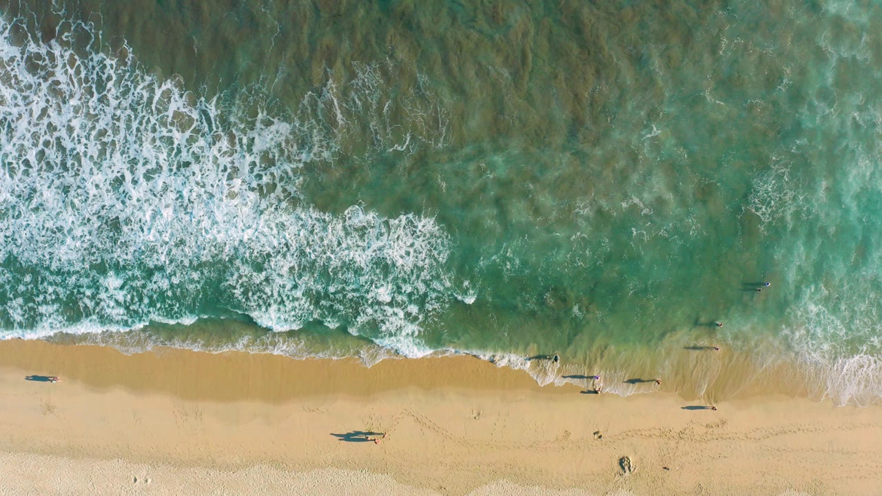 Overhead view in San Jose Del Cabo, Mexico, with white surf spreading across turquoise water toward a tan sandy beach and no visible people.