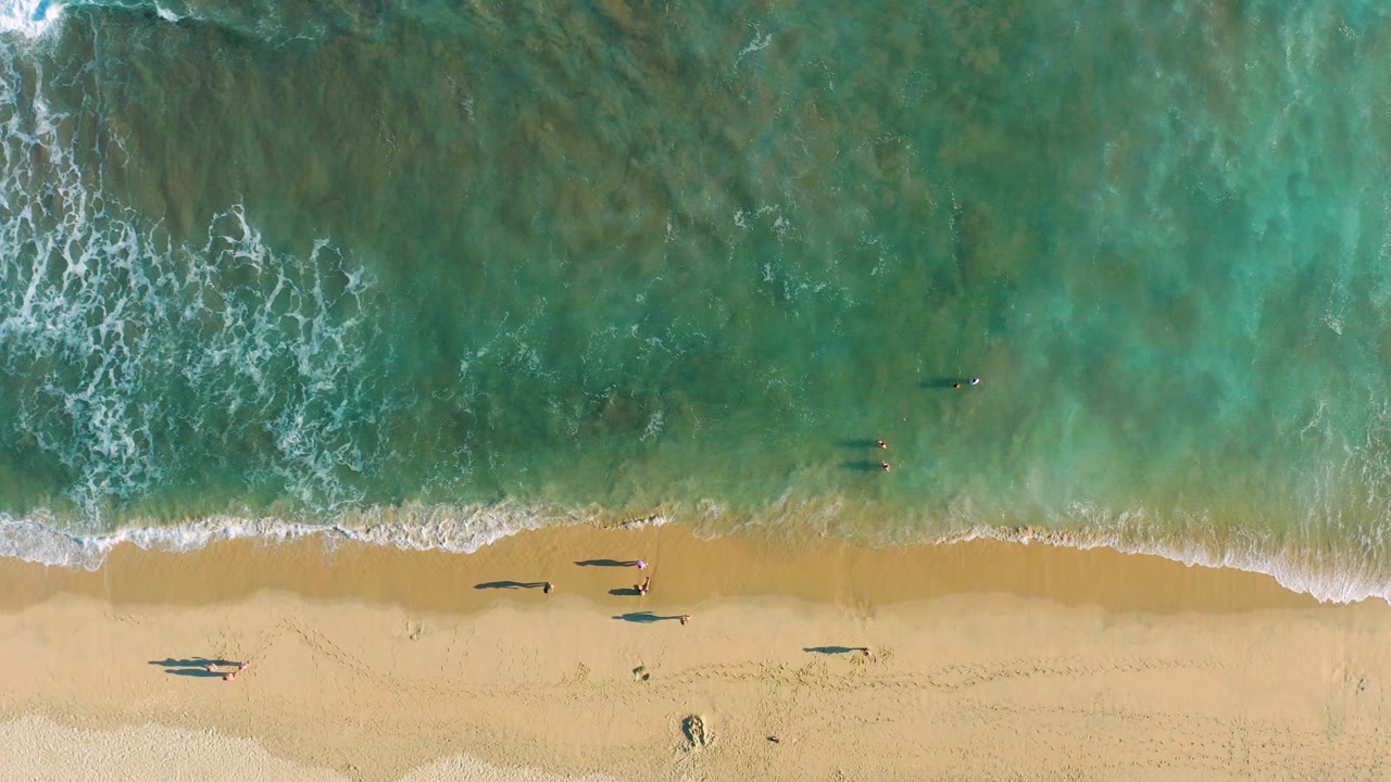 Overhead view of a San Jose del Cabo beach at the start of the clip, with turquoise surf entering from the top, pale sand below, and small beachgoers and surfers near the waterline.