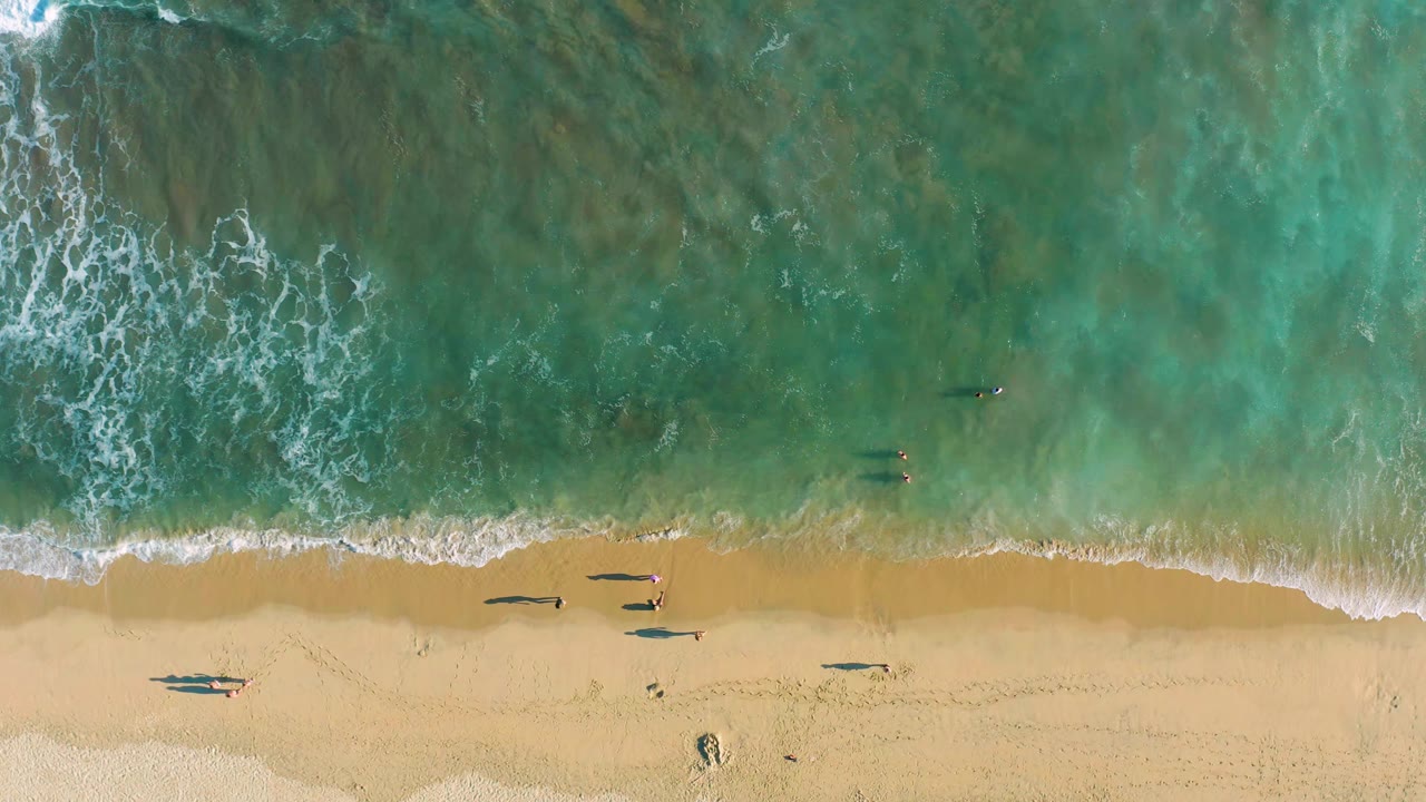 Aerial shoreline with white foam reaching the sand and people spaced along the water.