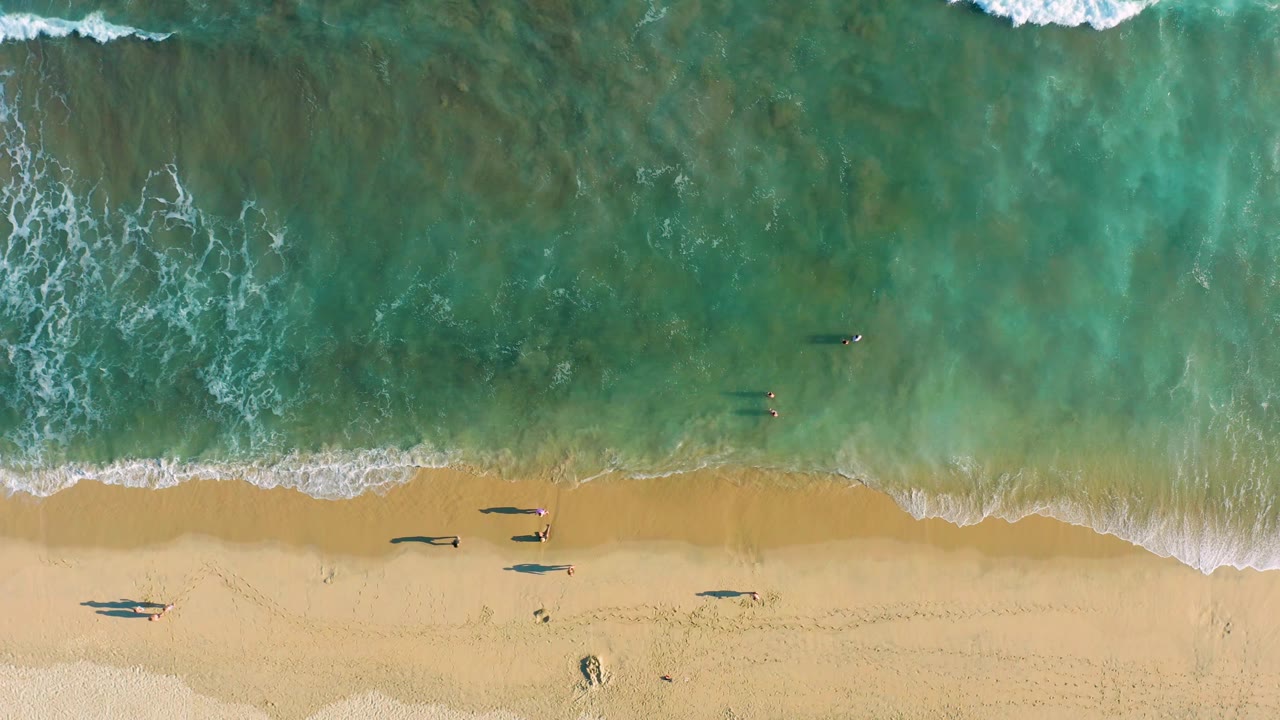 Top-down San Jose del Cabo beach frame with textured turquoise water above the shore, small surfers or swimmers in the water, and beachgoers casting shadows on the sand.