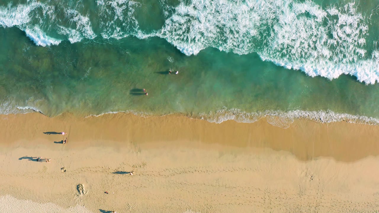 Aerial shoreline with white waves, footprints, and people scattered on sand and in water.