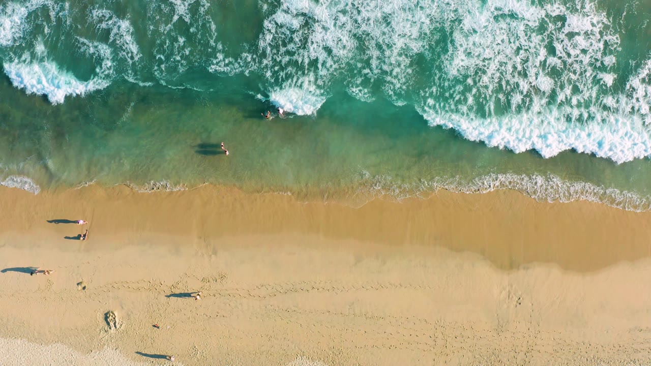 This San Jose del Cabo overhead frame shows foamy surf near the top, wet sand below it, and small people casting long shadows along the beach edge.