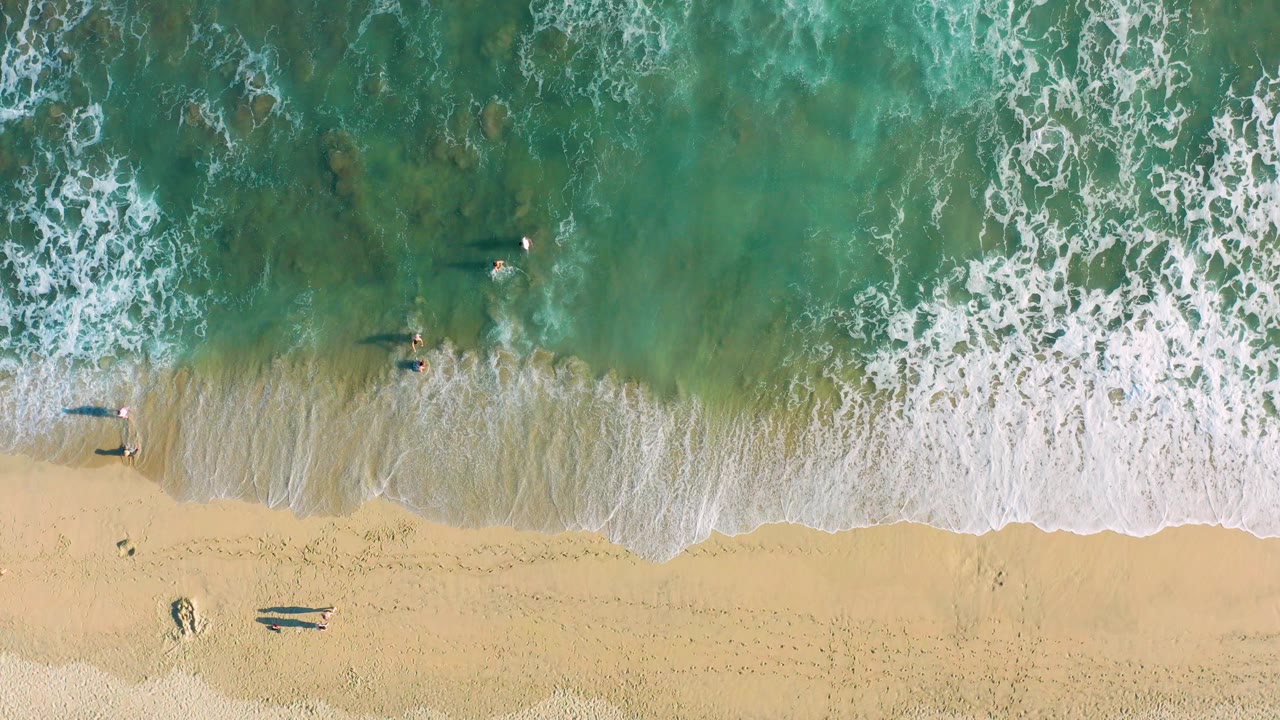 Overhead view of San Jose del Cabo beach where green-blue water breaks into white foam over tan sand with a few small people visible near the shoreline.
