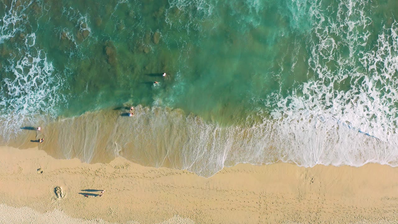 Top-down San Jose del Cabo beach frame with turquoise water, mottled darker patches below the surface, a swimmer near the break, and two small people on the sand.