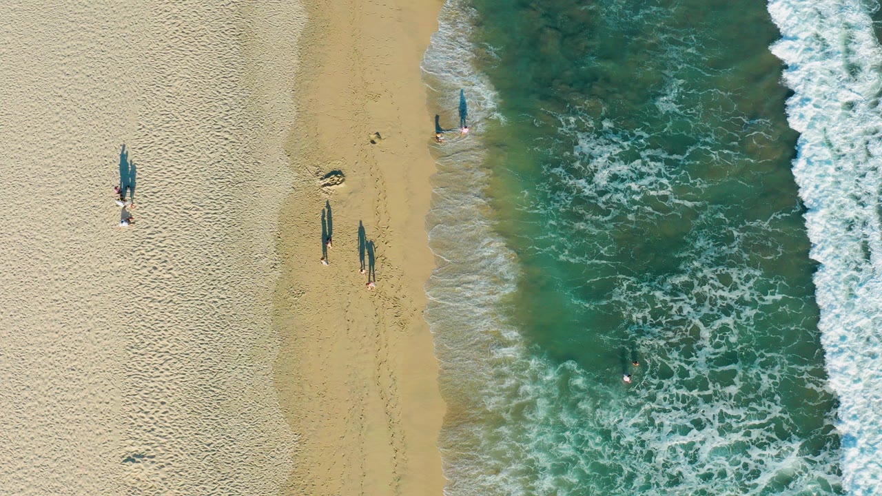 Overhead view in San Jose Del Cabo, Baja California Sur, with pale sand at left, green-blue surf at right, several visible beachgoers near the shoreline, and a swimmer offshore.