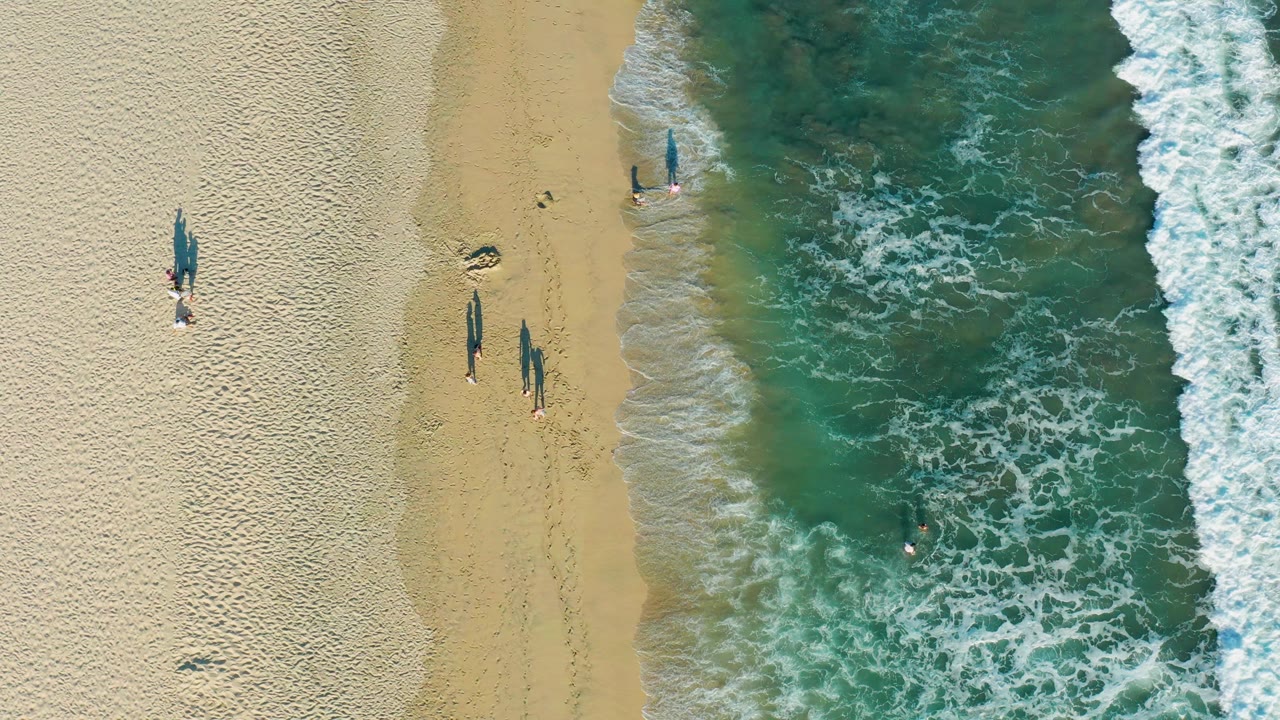 Aerial shoreline with walkers on sand and waves along the right edge.