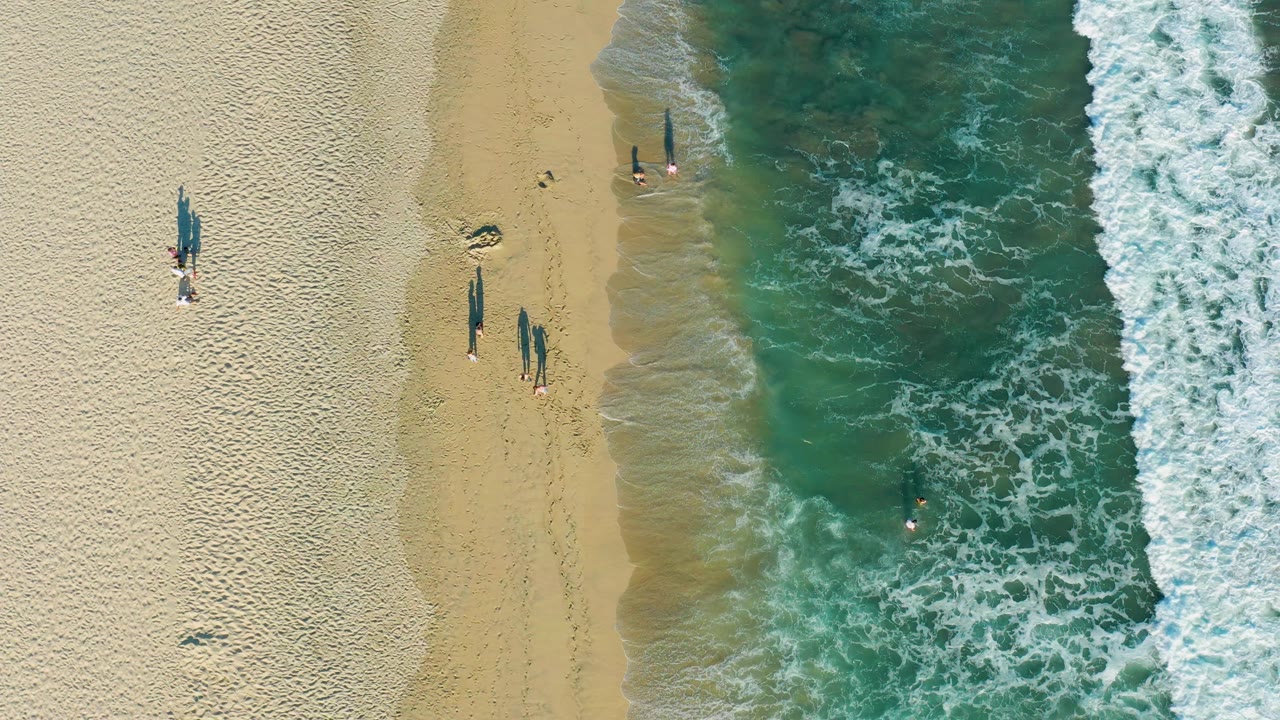 Top-down San Jose Del Cabo beach frame with small groups of visible people along the sand, a swimmer in the water, and textured white surf moving in from the right.