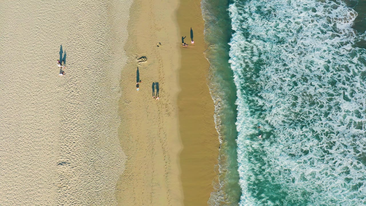 Overhead view in San Jose Del Cabo shows pale sand, a narrow wet shoreline, foamy turquoise surf, and several small beachgoers casting long shadows.