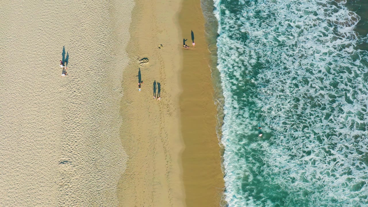 Overhead San Jose Del Cabo view shows the surf line slightly farther across the right side, with beachgoers on the sand and one small figure near the water.