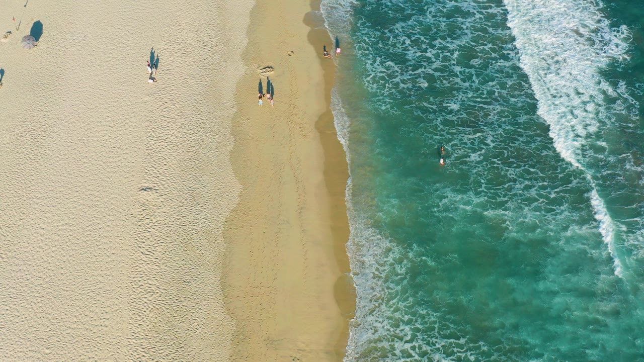 Overhead frame from San Jose del Cabo shows pale sand on the left, turquoise water on the right, white foam along the shore, and several small beachgoers near the waterline.
