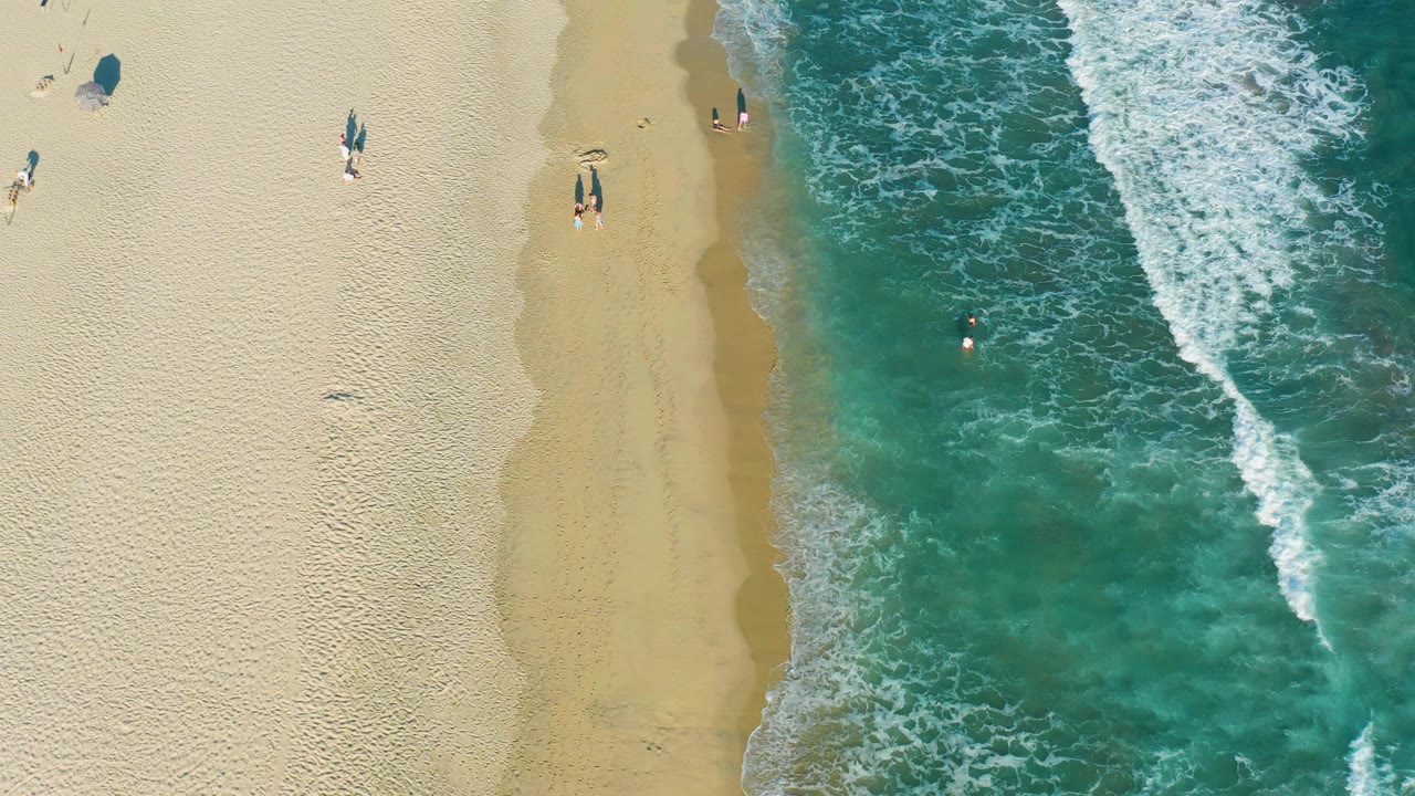 At 0:02, the overhead San Jose del Cabo view shows a smoother band of wet sand, white wave texture on the right, and small beachgoers separated across the upper sand and waterline.