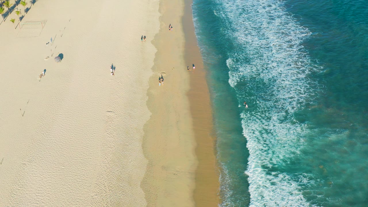 Overhead view of a San Jose Del Cabo beach with pale sand on the left, turquoise water on the right, white surf at the shoreline, and small people walking or standing near the water.