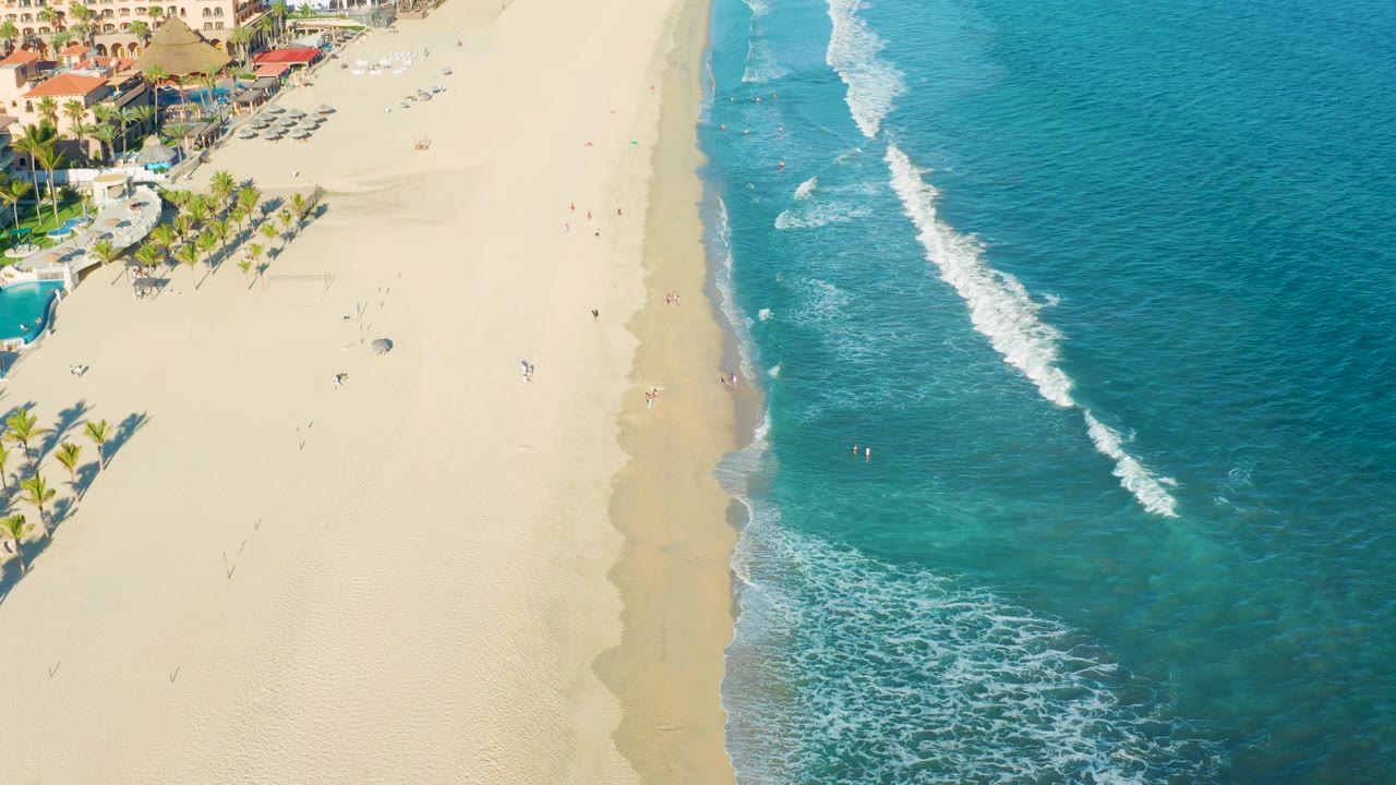 San Jose del Cabo aerial beach frame with more open ocean visible on the right, a foamy wave line near shore, and resort-area buildings and palms at the upper left.