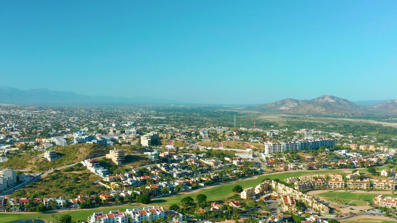 Wide aerial view over San Jose Del Cabo with rooftops, green open space, low-rise buildings, hazy mountains, clear blue sky, and no visible people.