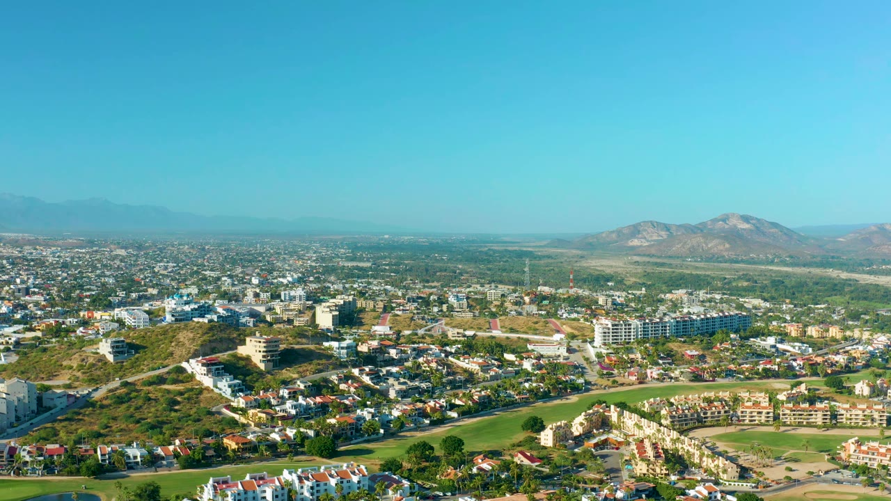 San Jose Del Cabo aerial frame showing foreground homes, a broad green corridor, midground urban buildings, distant mountains, bright daylight, and no visible people.
